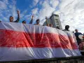 Ludzie trzymają biało-czerwoną flagę, symbol wolności Białorusi. Widać budynek w tle.