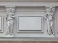 Dwie kamienne figury kobiet podtrzymują ozdobny gzyms. Jedna trzyma pędzel i paletę, druga opiera rękę na głowie.
