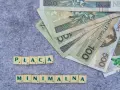 Płaca minimalna w Polsce – polskie banknoty o nominałach 100 i 500 zł ułożone na szarym tle, obok kafelki z literami tworzące napis "PŁACA MINIMALNA".