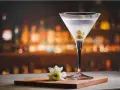 Eleganckie martini z colą, ozdobione oliwkami, na tle rozmytego baru. Obok kieliszka leży biały kwiatek.