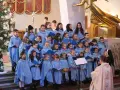 Dzieci w błękitnych strojach tworzą schola, śpiewając w kościele przy choince.