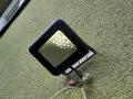 Czarna lampa led żarzy się po włączeniu, oświetlając zieloną, chropowatą ścianę. Widać przewody i złączki.