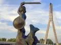 Syrenka Warszawska z mieczem i tarczą, symbol legendy o syrence warszawskiej, na tle nowoczesnego mostu.