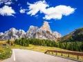 Strada panoramica per esplorare i passi del Trentino in moto, con imponenti vette dolomitiche sullo sfondo e un cielo azzurro con nuvole bianche.