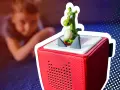 Ein grüner Drache mit Ei auf einer roten Toniebox, die blau blinkt. Ein Kind liegt unscharf im Hintergrund.