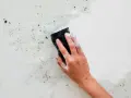 Main d'une personne lavant un mur humide avec une éponge savonneuse. Des saletés et des débris sont visibles sur la surface.
