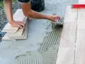 Układanie płytek na betonowym tarasie. Ręce pracownika rozprowadzają klej, obok stos płytek i poziomica.