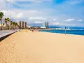 Słoneczna plaża w Barcelonie, palmy, deptak i błękitne morze. W oddali widać panoramę miasta.