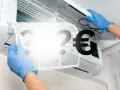 Hände in blauen Handschuhen entfernen einen Klimaanlagenfilter. Fragezeichen und ein Euro-Symbol deuten auf die **Klimaanlage reparieren Kosten** hin.