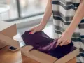 Kobieta pakuje ciemnofioletową sukienkę do kartonu, przygotowując ją do **vinted zwroty**.