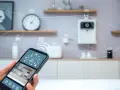 En hand håller en smartphone med en smart life app. I bakgrunden syns ett kök med uppkopplade enheter.