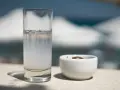 Vaso de pastis bebida, con una aceituna en un cuenco blanco. El fondo está desenfocado, sugiriendo un día soleado.