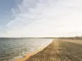 Spokojny brzeg jeziora Białe Kaszuby, fale delikatnie muskają piaszczysty brzeg, a na horyzoncie widać zarys zabudowań.