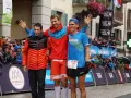 Trzech biegaczy na podium po ultra-trail du mont-blanc. Jeden w pomarańczowej kurtce, drugi w czerwonej, trzeci w niebieskiej koszulce.