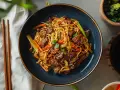 Pyszny makaron chow mein z wołowiną i warzywami, posypany sezamem. Obok pałeczki i miseczka z sosem sojowym.