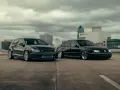 Dwa czarne samochody, sportowy hatchback i obniżony sedan, stoją na parkingu z widokiem na miasto.