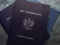 Stos dokumentów, na wierzchu ciemnogranatowy akt notarialny z godłem Polski.