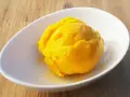Ein leuchtend gelber Löffel Mango Sorbet in einer weißen Schale auf Holzhintergrund.