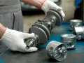 Mechanik w białych rękawiczkach trzyma wał korbowy, obok leżą tłoki. Uszczelniacz na wale jest kluczowy dla silnika.