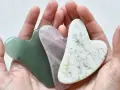 Drei herzförmige Gua Sha Steine in Grün, Rosa und Weiß liegen in einer Hand. Ideal für Gua Sha gegen Falten.