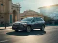 Ciemnoszary Toyota Corolla Cross Hybrid sunie ulicą miasta, odbijając światło słoneczne od nowoczesnych budynków.