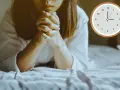 A woman is praying at 3am in the morning, her hands clasped tightly. The clock shows the early hour.