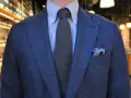 Elegancki mężczyzna prezentuje nietypowe wiązanie krawata. Szary, dzianinowy krawat idealnie komponuje się z niebieską marynarką i wzorzystą poszetką.