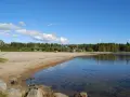 Solig strand vid Nydala camping Umeå med klart vatten och grönskande skog i bakgrunden.