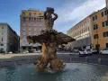 Fontana del Tritone in Piazza Barberini, Roma, con edifici storici e traffico sulla via dei Cappuccini.