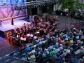 Eine Brass Band spielt auf einer Bühne, während ein Publikum auf Stühlen sitzt und zuhört.