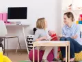 Terapeutka pracuje z dziewczynką, wspierając ją w terapii afazji u dzieci. W tle widać pomoce dydaktyczne i zabawki.