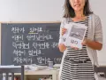 Lehrerin hält Bücher für einen koreanisch sprachkurs. Auf der Tafel stehen Sätze auf Koreanisch.