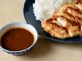Chrupiący kotlet schabowy polany sosem tonkatsu, obok biały ryż i miseczka sosu.