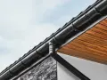 Czarny system rynnowy odprowadza wodę opadową z dachu. Widoczny fragment elewacji z kamienia i drewnianego podsufitki.