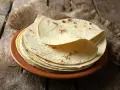 Stos cienkich, jasnych placków keto tortilla na brązowym talerzu, ułożonych na płótnie i drewnianym blacie.