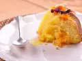 Un trozo de bizcocho de almendra de la abuela, decorado con ralladura de naranja y flores secas, servido en un plato blanco con una cucharilla.