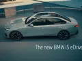 Jasnoszary sedan BMW i5 eDrive40 stoi na zewnątrz, z widokiem na basen i ławkę.