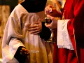 A priest in red vestments holds a chalice and host, while another in white vestments holds a paten. A moment of communion, perhaps a prayer to St. Benedict for healing.