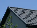 Szary dach z blachy, idealny do obliczeń, jak obliczyć blachę na dach. Widać fragment ściany i gałęzie drzew.