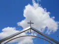 Ein Kreuz ragt über einem modernen Torbogen in den blauen Himmel mit Wolken. Ein starkes Christi Himmelfahrt Symbol.