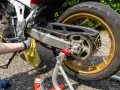 Mano guantata pulisce la catena della moto con uno straccio giallo. Spray per la pulizia catena visibile.