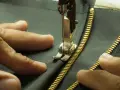 Main tenant une fermeture éclair dorée, une aiguille de machine à coudre s'apprête à coudre une fermeture éclair à la main sur un tissu sombre.