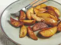Goldbraune Kartoffelspalten, frisch aus dem Ofen, liegen auf einem Teller. Ein Messer und eine Gabel liegen daneben.