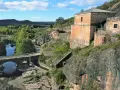 El portón de la oliva, un antiguo edificio de piedra junto a un río y un puente de arco, se alza sobre un paisaje rocoso y verde.