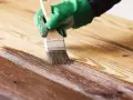 Mano guantata applica vernice protettiva con pennello per impermeabilizzare il legno, creando una finitura lucida.