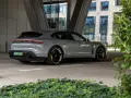 Szare Porsche Taycan Turbo S stoi pod zadaszeniem, z widokiem na nowoczesny budynek i zieleń.