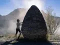 Une femme interagit avec une structure ovoïde en pierres, une œuvre de land art, dans un paysage brumeux et montagneux au lever du soleil.
