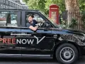 Ein Mann fährt ein schwarzes freenow taxi. Im Hintergrund steht eine rote Telefonzelle.