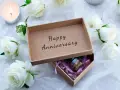 Otwarta szkatułka z napisem "Happy Anniversary" i małą buteleczką w środku, otoczona białymi różami i płonącą świecą. Idealny prezent na 21 rocznicę ślubu.