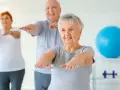 Trois seniors souriants pratiquent la gym douce, bras tendus, dans une salle lumineuse avec un ballon bleu en arrière-plan.